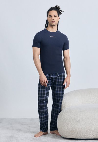 Άντρας με μακριά dreadlocks στέκεται ξυπόλυτος φορώντας ένα στενό μπλε ναυτικό T-shirt και μπλε καρό πυτζάμες δίπλα σε έναν άσπρο υφασμάτινο καναπέ.