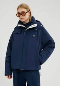 Granatowa zimowa kurtka z odpinanym kapturem, białymi akcentami i dwoma przednimi kieszeniami. Wykonana z gładkiego, wodoodpornego materiału.
