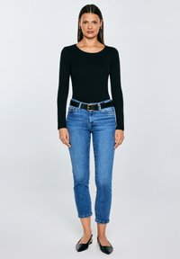 Naine seisab, kandes musta pika varrukaga pikka t-särki, siniseid lühikesi teksapükse musta vööga ja musti terava ninaga slingback-platvormkingi neutraalsel taustal.