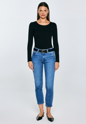 Sieviete stāv, valkājot melnu garroku topu, zilas īsās džinsa bikses ar melnu jostu un melnus punktainus sandaļus ar atvērtu papēdi uz neitrālas fona.