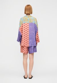 Άτομο με κοντά κόκκινα μαλλιά που φορά φαρδιά πουκάμισο με πολλαπλά σχέδια και σορτς σε παστέλ αποχρώσεις μπλε, μωβ και ροζ, στέκεται με την πλάτη προς την κάμερα.