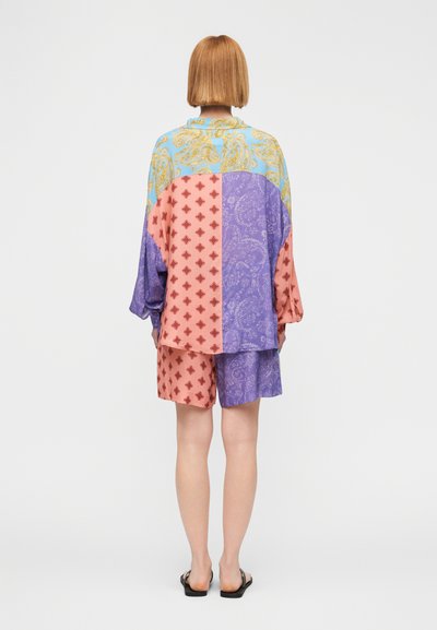 Άτομο με κοντά κόκκινα μαλλιά που φορά φαρδιά πουκάμισο με πολλαπλά σχέδια και σορτς σε παστέλ αποχρώσεις μπλε, μωβ και ροζ, στέκεται με την πλάτη προς την κάμερα.