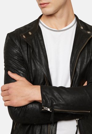 Black leather jacket with a textured finish, silver hardware, zip details, and a standing collar, worn over a white T-shirt.