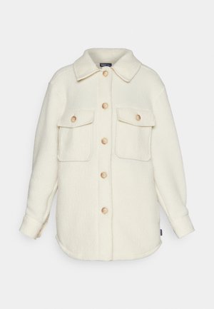 Chaqueta de camisa de forro polar color crema, con cuello, mangas largas y dos bolsillos frontales. Presenta botones de madera clara y un dobladillo redondeado.