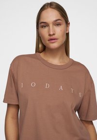 T-shirt in cotone marrone con maniche corte e scollatura tonda, che mostra la scritta bianca "10 GIORNI" sul fronte. Texture liscia.