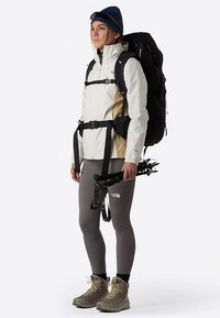 Biało-beżowa kurtka przeciwwiatrowa z czarnymi akcentami, szare legginsy i beżowe buty trekkingowe. Nosi duży czarny plecak i kijki trekkingowe.