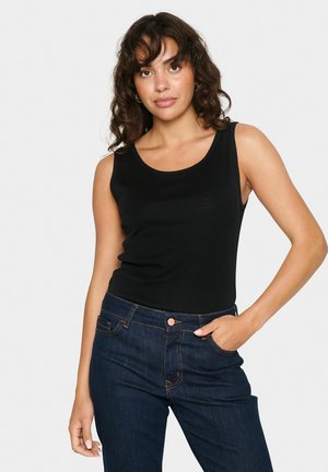 Débardeur noir sans manches avec un col rond, fabriqué en tissu lisse. Porté avec un jean en denim foncé affichant des détails de couture visibles.
