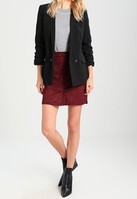 Blazer noir porté sur un haut gris, associé à une jupe en daim bordeaux. Des bottines noires complètent la tenue, mettant en valeur une silhouette ajustée.