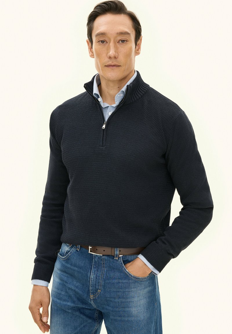 Granatowy sweter na zamek z fakturowanym wzorem i ściągaczem przy kołnierzu, noszony na jasnoniebieskiej koszuli, w połączeniu z niebieskimi dżinsami.