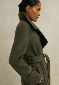 Olive roheline villane mantel, millel on sisselõigatud krae, küljetaskud ja vöö taljes. Tekstuuriga kangas ja nööbid.