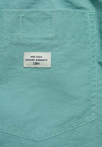 Látka v mätovej zelenej farbe s textúrovaným povrchom. Obsahuje našitú etiketu s nápisom "HNG TEEN GENUINE GARMENTS 1984" nad vreckom.