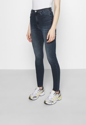 Persona che indossa jeans skinny scuri e sneakers bianche con dettagli gialli e grigi, in piedi davanti a uno sfondo chiaro e uniforme.