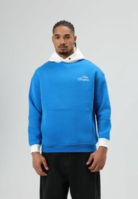 Felle blauwe trui met een witte capuchon, voorzien van een voorzak en een geborduurd logo. Zachte stof met geribbelde manchetten en zoom.