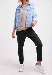 Hellblaue Jeansjacke mit Used-Optik, beiger Pullover, schwarze Hose und weiße Sneaker mit blauen Akzenten.