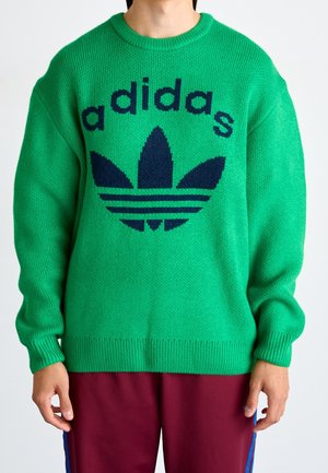 Personne portant un pull en tricot vert vif avec un grand logo Adidas bleu marine et un pantalon bordeaux avec des rayures bleues.
