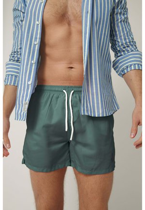 Hombre con camisa de rayas azules y blancas desabrochada, con las mangas remangadas y pantalones cortos de baño verdes con cordón blanco.