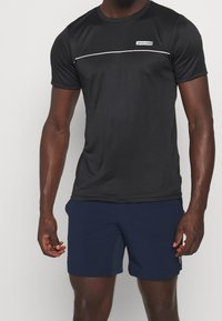 Hombre con una camiseta atlética negra de manga corta con ribete blanco y pantalones cortos azul marino, de pie contra un fondo claro y liso.