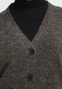 Hnědý pletený kardigan s designem do V, s dvěma tmavými knoflíky a měkkou, texturovanou látkou. Nosený přes černý rolák.