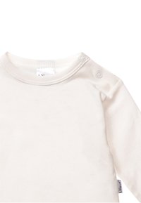 Helle beige Langarmshirt aus weich Baumwollstoff. Verfügt über einen Rundhalsausschnitt mit zwei Druckknöpfen an der Schulter und einem kleinen Etikett an der Seite.