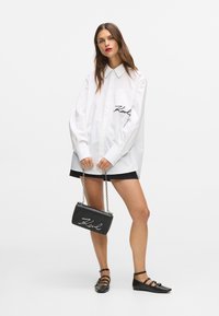 Hvit oversized skjorte med svart logo, sammen med et svart miniskjørt, svart håndveske med kjede-stropp, og svarte flate sko med stropper.