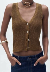 Brązowy dziergany kamizelka z głębokim dekoltem w kształcie litery V, z przodu z guzikami i subtelną teksturą. W połączeniu z ciemnoniebieskimi dżinsami i złotą bransoletką.