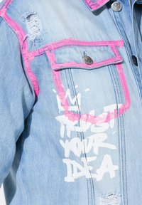 Veste en denim bleu clair avec des zones déchirées, des accents rose vif et du texte blanc qui dit "JE FAIS CONFIANCE À VOTRE IDÉE."