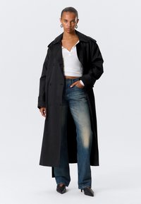 Cappotto lungo nero con design doppiopetto, abbinato a jeans a gamba larga azzurri e a un top bianco aderente. Scarpe nere a punta.
