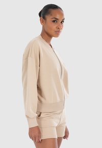 Veste bomber beige zippée avec des poignets et un ourlet côtelés, dotée d'une coupe décontractée et d'un tissu doux. Portée avec un short assorti.