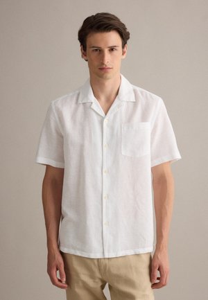 Giovane uomo che indossa una camicia bianca a maniche corte e pantaloni beige, in piedi su uno sfondo semplice e neutro.