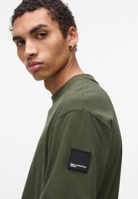 Tricou cu mânecă lungă de culoare verde măsliniu, fabricat dintr-un material moale, având un patch cu logo pătrat negru pe mâneca stângă și un guler rotund.