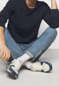 Marinblå stickad tröja, ljusblå denimjeans, vita strumpor och atletiska sneakers med svarta och blå accenter. Modell sitter med benen i kors.