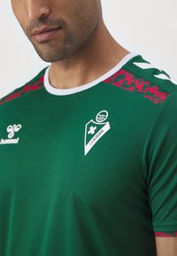 Zielona koszulka sportowa z krótkimi rękawami, biała kołnierzyk, czerwone wzorzyste akcenty na ramionach, logo Hummel oraz herb drużyny na klatce piersiowej.