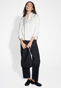 Camicia bianca con maniche arrotolate, abbinata a pantaloni neri a gamba larga e una borsa nera dalla texture. Modello in piedi su uno sfondo bianco.