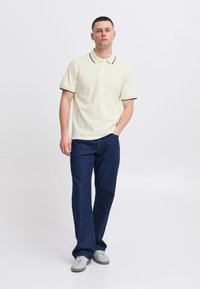 Polo skjorte i lys beige med mørkegrønn kant, sammen med mørkeblå jeans. Antrekket har en klassisk passform og en uformell stil.