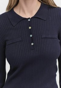 Marineblauw geribd poloshirt met een kraag, voorzien van een vijfknops sluiting en twee borstzakken. Zachte gestructureerde stof.