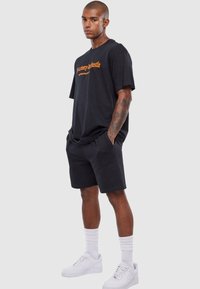 Black cotton t-shirt with orange embroidered text, paired with black shorts. White sneakers and crew-length white socks complete the outfit.