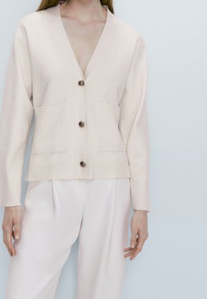 Light beige cardigan with a V-neck, long sleeves, two front pockets, and three dark buttons. Worn over white trousers. Smooth texture.