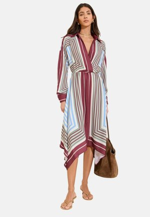 Frau trägt ein langärmeliges Midikleid mit V-Ausschnitt und geometrischen Streifen in Burgunderrot, Blau und Weiß, hält eine braune Wildledertasche und trägt Sandalen.