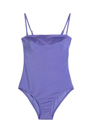 Kvinde med mellemtonet hudfarve iført en lilla badedragt i ét stykke, poserer med en arm, der dækker en del af hendes ansigt, og smiler mod en hvid baggrund.