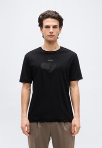 Νέος άνδρας που φορά μαύρο μπλουζάκι με το λογότυπο BOSS και μπεζ παντελόνι, στέκεται μπροστά σε ένα απλό ανοιχτό γκρι φόντο, κοιτάζοντας μπροστά.