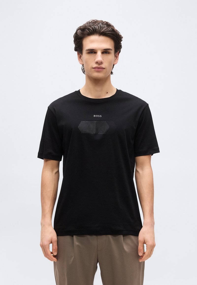 Νέος άνδρας που φορά μαύρο μπλουζάκι με το λογότυπο BOSS και μπεζ παντελόνι, στέκεται μπροστά σε ένα απλό ανοιχτό γκρι φόντο, κοιτάζοντας μπροστά.