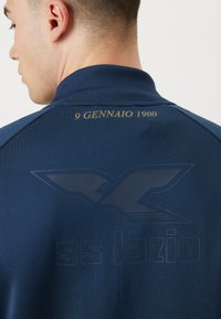 Veste de sport bleu marine avec col haut, arborant un logo en relief et le texte "9 GENNAIO 1900" en doré au dos. Texture lisse.