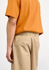 Oranssi puuvillapaita yhdistettynä beigeihin housuihin. Housuissa on yksi takatasku napilla ja ne ovat sileää, teksturoitua kangasta.