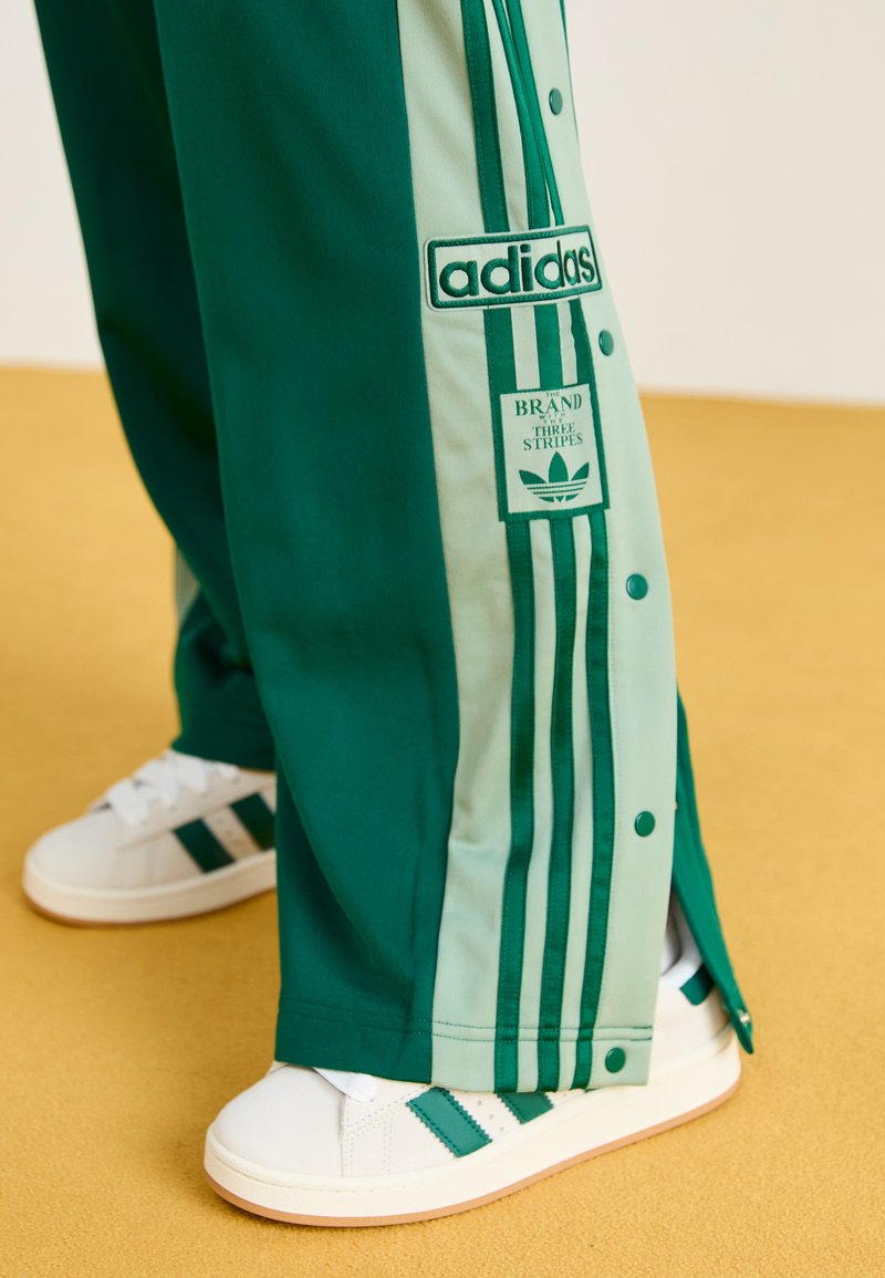 Pantalones deportivos verdes con paneles laterales verde menta y detalles de tres rayas. Presentan botones a presión y un parche de marca. Combinados con zapatillas blancas.