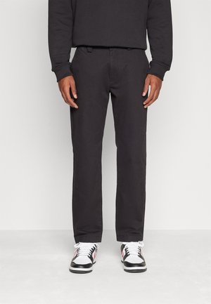 Pantalones chinos negros de algodón con un corte recto, que cuentan con bolsillos laterales y una textura suave, combinados con zapatillas negras y blancas.