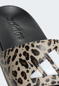 Klapki Adidas z beżowym paskiem w panterkę, białym logo z trzema paskami oraz teksturowaną czarną podeszwą. Płaski i szeroki fason.