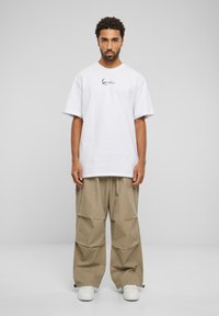 T-shirt blanc surdimensionné avec logo noir, associé à un pantalon large beige à design plissé, porté avec des baskets blanches.