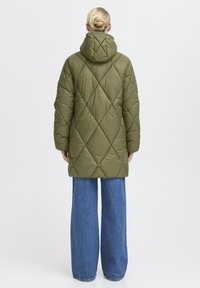 Olijfgroene gewatteerde jas met capuchon en een ruitvormig quiltpatroon, met een middellange snit en wijde mouwen. Draag je over lichtblauwe wijde jeans.