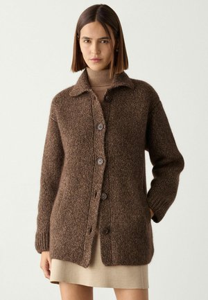 Kvinde iført en brun, knappet, strikket cardigan over en beige rullemekjole, stående mod en ensfarvet lys baggrund.