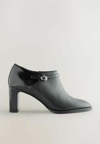 Botă ankle neagră cu toc, realizată din piele netedă, cu un accent lucios din patent, având o curea decorativă cu un cataramă argintie și vârf ascuțit.